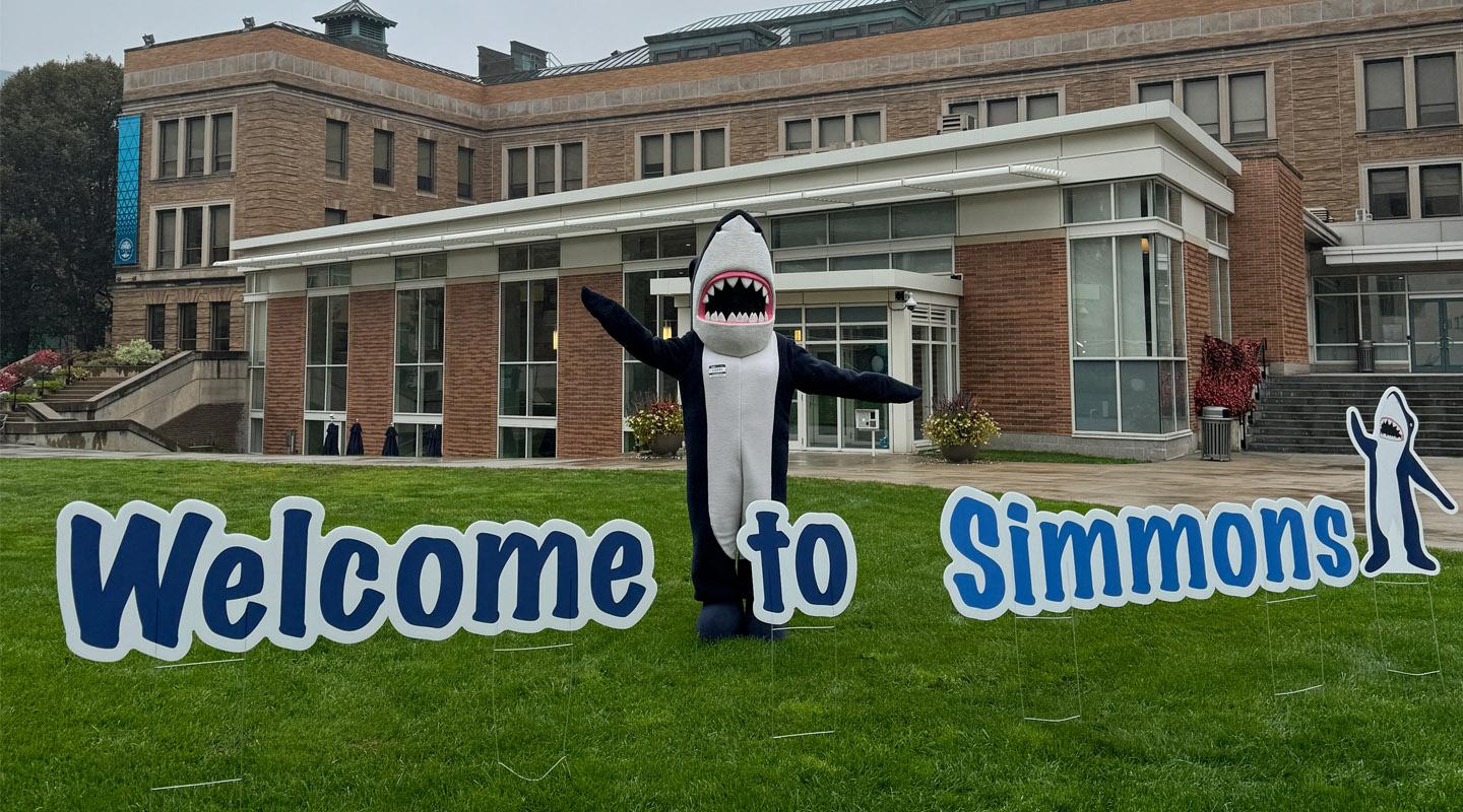 Welcome to Simmons! Stormy standing with welcome sign on lawn of main quad