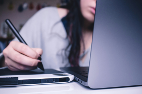 A student taking notes in a notebook while reading from a laptop