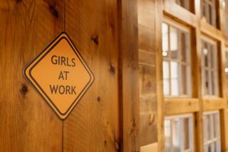 Girls at Work logo posted on a wall.  An orange, diamond shaped sign with the words "Girls at Work" in the center.