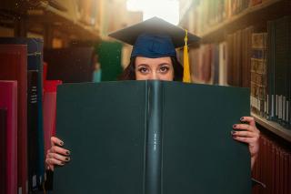 A student standing in library stacks wearing a grad cap and peering over the top of an open book