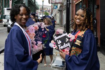 2 graduating students with decorated grad caps.  One cap says "She's Grad," and the other says "Someday is Today"