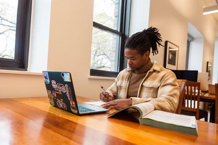 A student sits at a table in a library with an open book and a laptop