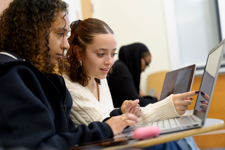 Two students in classroom desks looking at one laptop screen